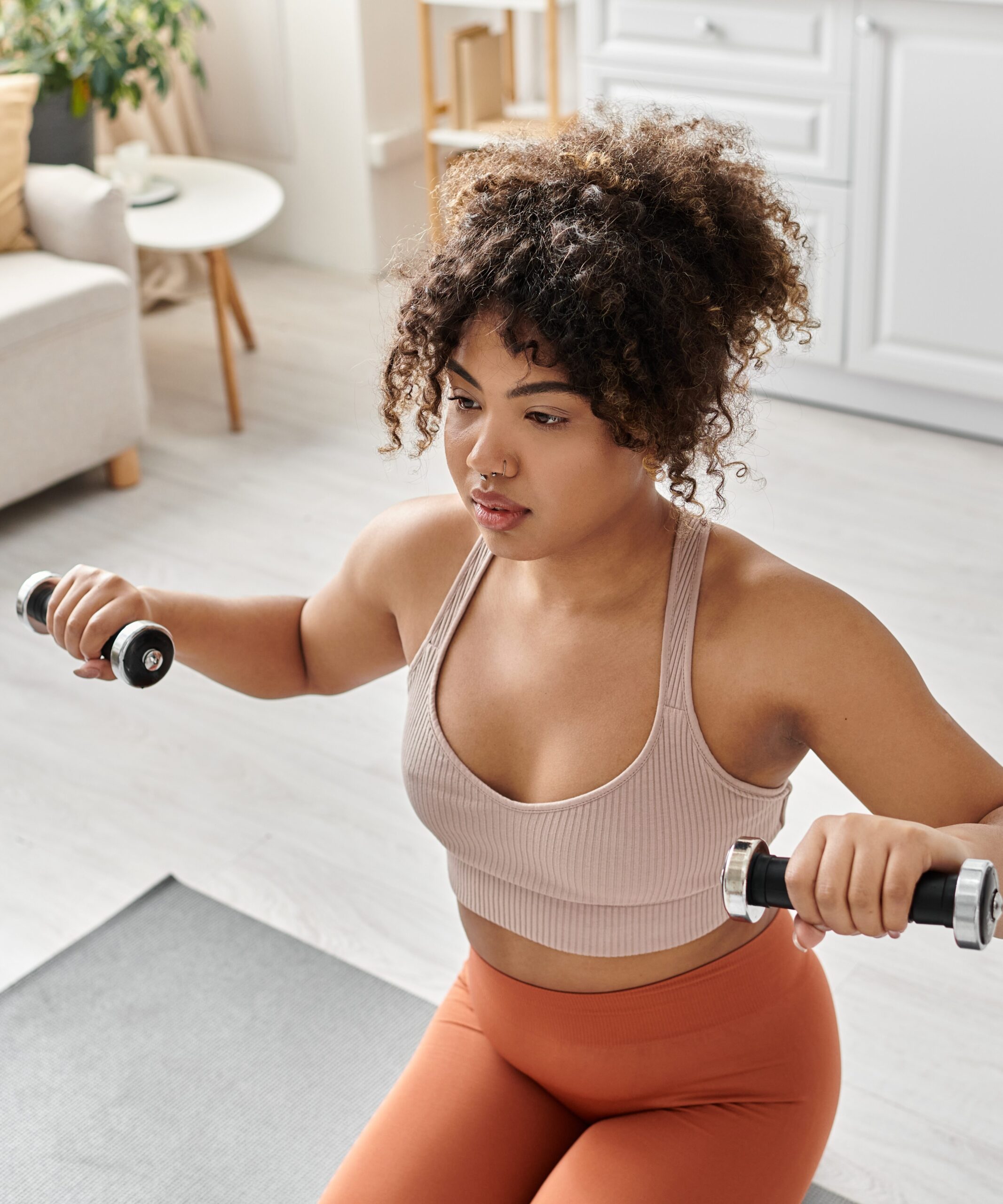 woman on yoga mat in her home holding dumbbells at shoulder height doing a workout 
